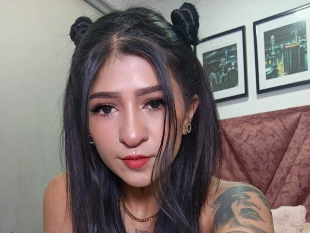 LuciadelaVega, con codini neri, piercing al naso e orecchini, ha dei tatuaggi, rossetto rosso e una collana a catena d'oro, e guarda la fotocamera con un leggero sorriso..