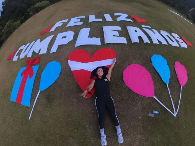 JessicaFiore lying on grass, arms out, wearing black outfit and white sneakers, with colorful heart, balloons, and text on ground, looking happy, tattoos visible on her arm.