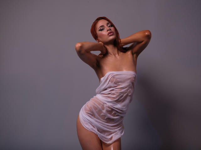 JesicaRare with red hair, wearing a wet, sheer white dress, exposing her tits and pussy, hands behind head, gray background, seductive pose, medium shot, soft lighting.