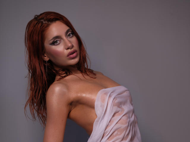 JesicaRare with wet red hair, glistening skin, and a sheer pink cloth covering her tits, looking seductively at the camera. She's wearing dark eyeliner and subtle pink lipstick.