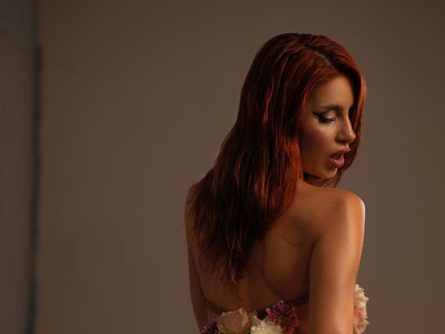 JesicaRare with red hair, looking over her shoulder, wearing floral straps covering her tits, in soft brown lighting, showing her bare back and slightly parted lips.