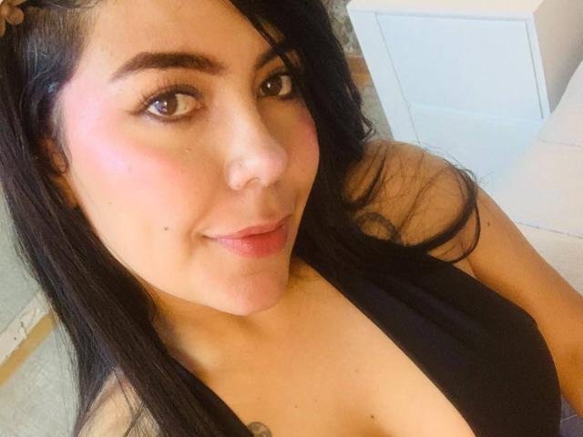 CatalinaReiyna with black hair, brown eyes, pink blush, subtle smile, black top, visible cleavage, small tattoo near collarbone, light skin, cute face, medium-sized tits, bedroom setting.