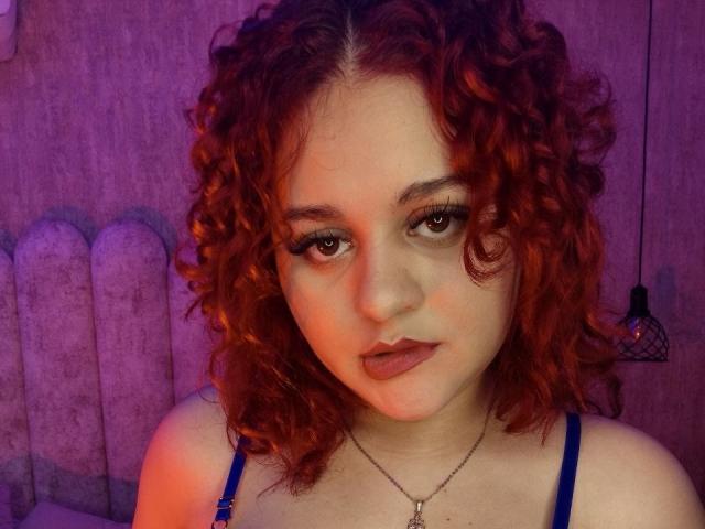RosalieVittoria with curly red hair, wearing a blue bra, has pouty lips and a necklace, staring with brown eyes, in a purple-lit room.