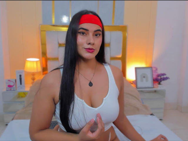 AlaraKey with long black hair, red headband, white tank top, and necklace, touching her tits in a bedroom with warm lighting.