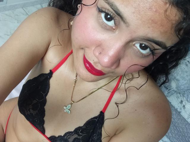 ValeryPoleth, con i capelli neri ricci, rossetto rosso, reggiseno di pizzo nero con spalline rosse, una collana dorata con un ciondolo a forma di farfalla, guarda verso l'alto, sorridendo leggermente, mostrando il suo piercing al naso e il suo viso carino..