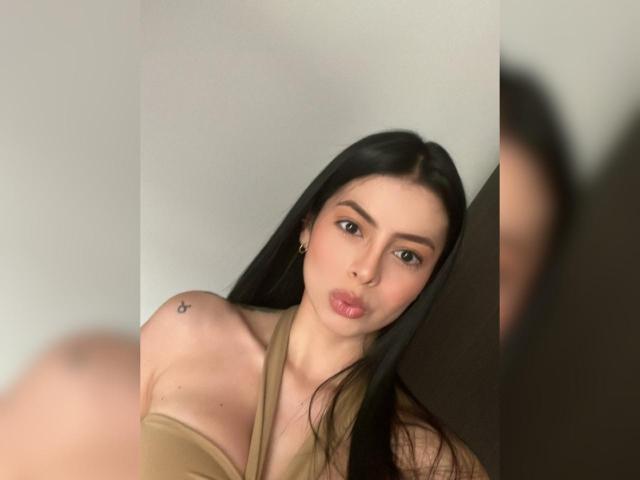 IsabellaJames with long black hair, pouty lips, small tattoo on right shoulder, and off-shoulder beige top, looking at camera. Her tits are slightly visible. Blurred reflection on right.