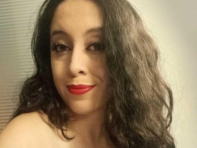 Kendra Simons, con i capelli neri ricci, il rossetto rosso e le lunghe ciglia, sorride alla fotocamera, mostrando le spalle scoperte. Ha un viso carino e un aspetto seducente..