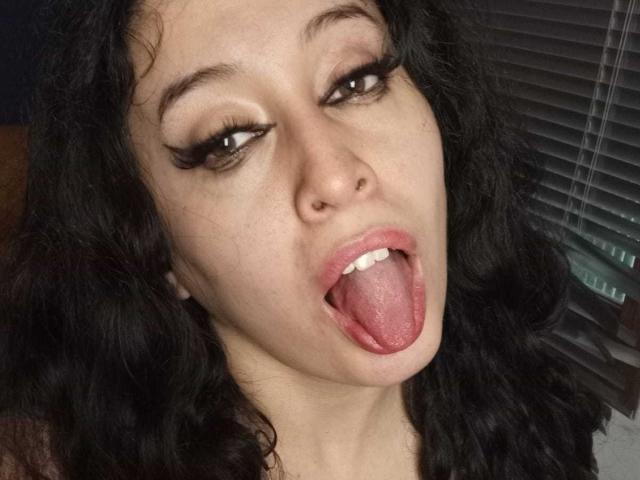 KendraSimons has curly black hair, brown eyes with heavy eyeliner, and is sticking out her pink tongue with her mouth slightly open. Her nose is pierced on the right.