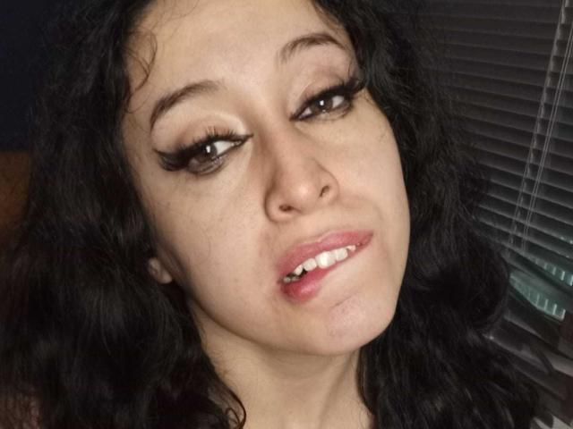Kendra Simons, con i capelli neri ondulati, gli occhi marroni e le lunghe ciglia, le labbra rosa leggermente schiuse e un piccolo piercing al naso, guarda verso l'alto con un sorriso rilassato e discreto..