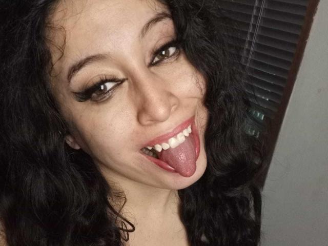 KendraSimons with curly black hair, brown eyes, and black eyeliner, sticking out her tongue and showing off her white teeth. Her tongue is visibly large and pink.