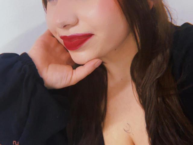 VenusxMusa with red lipstick, brown hair, and a black shirt, touching her cheek with a hand, showing a small tattoo near her cleavage. Her face is slightly blushed.