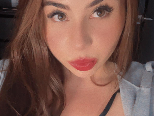 VenusxMusa has pouty red lips, long brown eyelashes, and light brown eyes. She's wearing a gray hoodie with black straps, brown hair, and a slightly pouty expression.