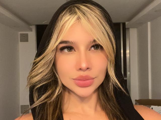 SashaPolet hat blondes und braunes, welliges Haar, trägt eine schwarze Kapuze, hat volle, rosafarbene Lippen, grüne Augen und einen leichten Schmollmund, auf einem Selfie..