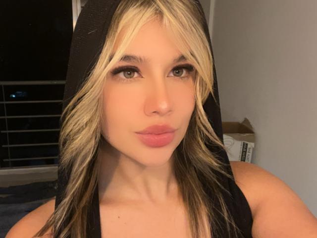 SashaPolet mit blonden und braunen Haaren, die eine schwarze Kapuze trägt, hat volle, rosafarbene Lippen und grüne Augen und blickt direkt in die Kamera, in einem schwach beleuchteten Raum..