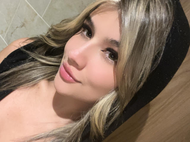 SashaPolet a de longs cheveux blonds, de grands yeux marrons et un joli sourire avec du rouge à lèvres rose. Elle porte un haut noir, et son visage est légèrement incliné vers la droite..