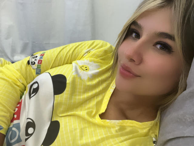 SashaPolet, avec ses cheveux blonds, portant un haut de pyjama jaune avec un motif panda, allongée, souriant légèrement, visage mignon, maquillage doux, petit nez, lèvres rosées discrètes, oreiller blanc en arrière-plan..