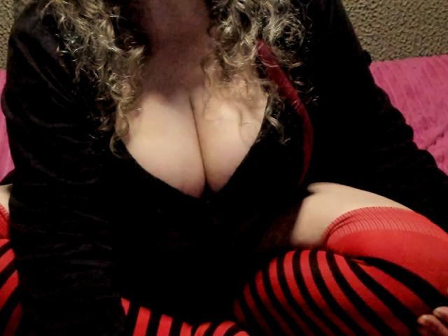 BeautyHairy with curly blonde hair, wearing a black top with a deep cleavage, showing her large, pale tits, and red striped thigh-high socks, sitting with legs crossed.