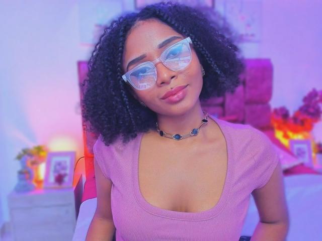 MeganWilliams with curly black hair, wearing clear glasses and a purple ribbed top, showing her small tits and pink glossy lips, wearing a black necklace with blue gems.