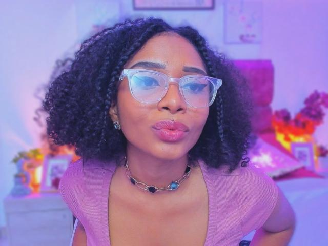 MeganWilliams with curly black hair, wearing transparent glasses and a pink top, has full pink lips and a necklace with blue and black stones. She's pouting slightly.