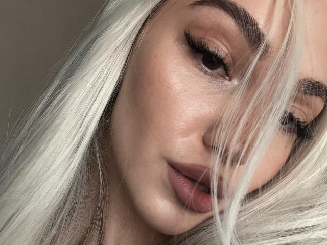SophieKitty's face with platinum blonde hair, thick eyebrows, long eyelashes, and full, pinkish lips. Her skin is smooth and slightly shiny. She looks down with a subtle, sexy expression.
