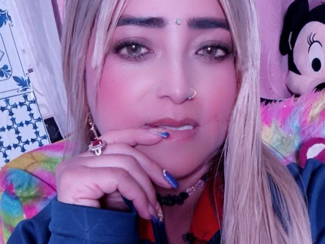 AlessadraJoness ha un leggero rossore sulle guance, smalto blu per le unghie, un piercing al naso e un copricapo. Capelli biondi, occhi marroni, una mano sul mento, indossa una camicia blu. Sfondo rosa e colorato..
