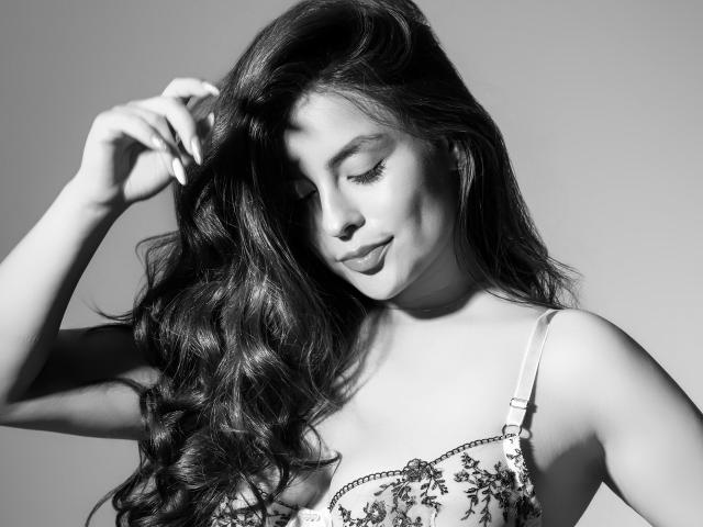 FerAram, avec de longs cheveux noirs ondulés, les yeux fermés, souriant doucement, portant un soutien-gorge à motifs floraux, une main dans les cheveux, photo en noir et blanc, montrant un visage doux, sexy, attrayant et mignon..