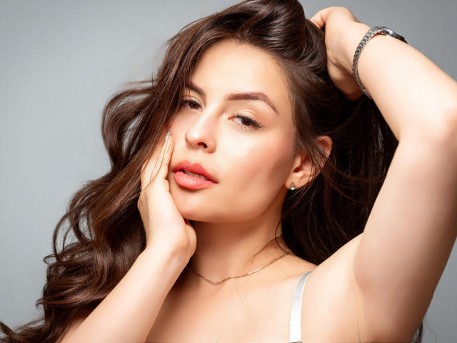 FerAram a les cheveux bruns ondulés, des lèvres pulpeuses et une peau claire. Elle touche sa joue avec une main, lève l'autre, porte un bracelet en argent et a un regard séduisant..