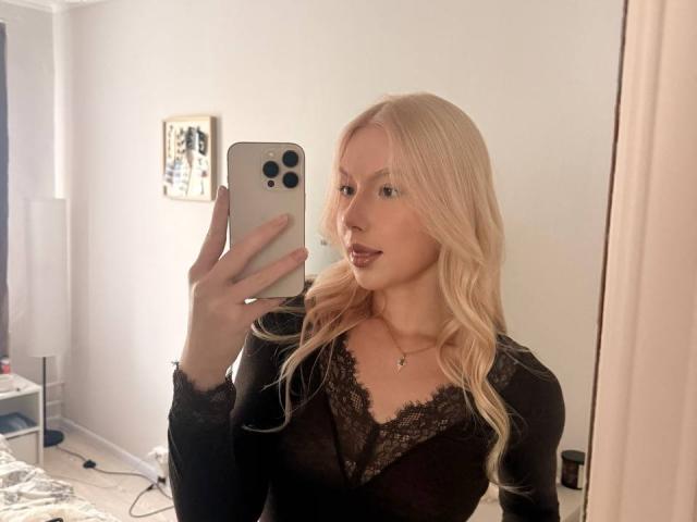 LaurindaLupacchino with blonde hair, wearing a black lace top, taking a mirror selfie with her iPhone, showing her tits and a subtle smile.