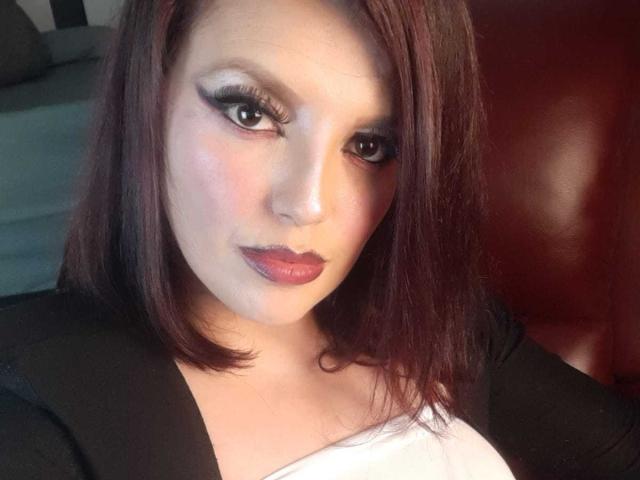 SachyMoon with shiny brown eyes, dark lipstick, and medium-length brown hair, wearing a black jacket over a white top, looking directly at the camera with a serious expression.