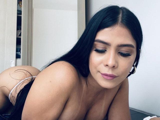 MademoiselleGo hat langes, schwarzes Haar, große Brüste und einen hübschen Hintern. Sie ist oben ohne, trägt weiße Kopfhörer, hat pinken Lippenstift auf und blickt mit einem leichten Lächeln nach unten..