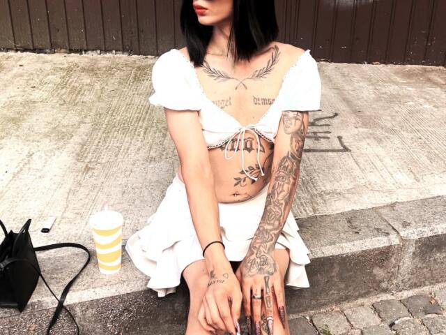 IrisIrs with black hair, white off-shoulder top, white skirt, and tattooed chest and arms, sitting on concrete, cup and bag to the left, showing her tits and ass.