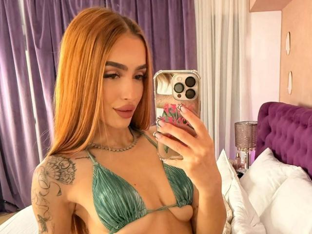 MarissaFex with long orange hair, green bikini top, tattoo on right shoulder, holding phone, smiling, showing small tits, in a bedroom, taking a selfie.