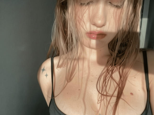 DelmyMasztal with wet, blonde hair covering her face, wearing a black top, showing small star tattoos on her right shoulder, and her puffy, pinkish-tan tits.
