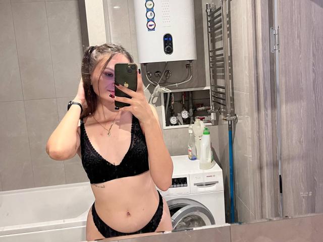 LoidaGess in a black velvet bra and panties, holding a phone in a bathroom mirror selfie, showing her tits and ass, with a washing machine and toiletries behind.