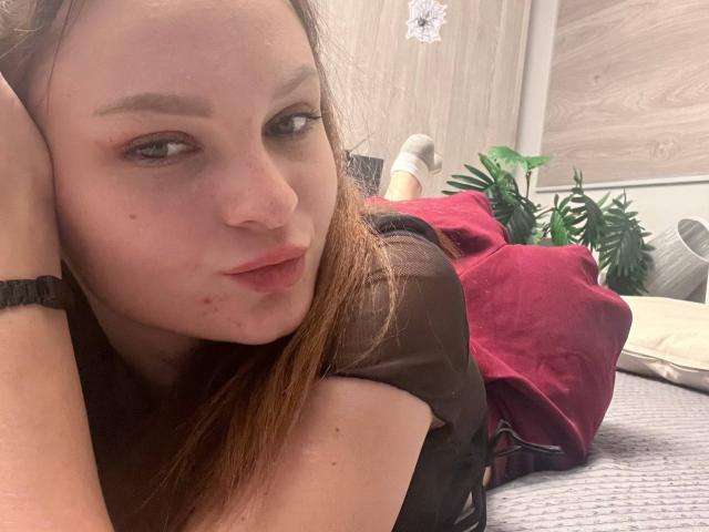 LoidaGess is lying on a bed, face up, with brown hair, wearing a black shirt, and a black bracelet on her wrist. Her ass is covered by a red blanket.
