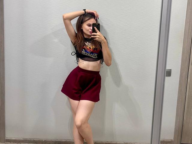 LoidaGess with long brown hair, small tits, wearing a black mesh crop top and red shorts, standing in a mirror selfie, right arm raised, pouty expression, holding phone.