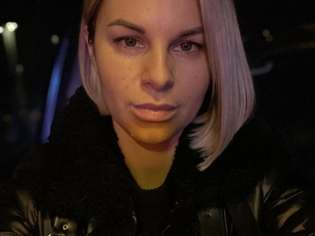 BelleLisaG with pale skin, short blonde bob, and serious expression, wearing a black leather jacket with a furry collar, in a dimly lit, dark background.