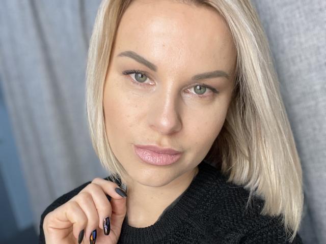 BelleLisaG with blonde hair, green eyes, and pink lips, wearing a black sweater, holding her collar with black nails. Her skin is light and she's looking at the camera.