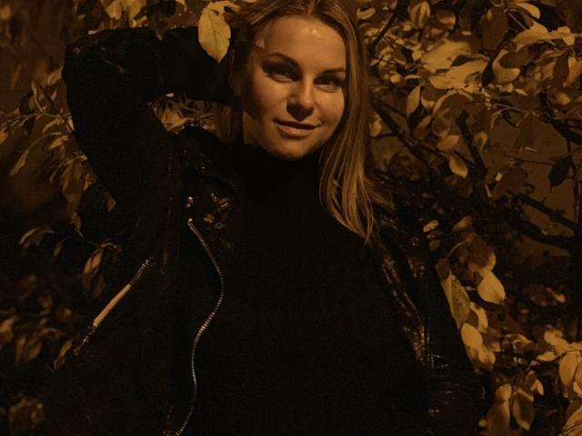 BelleLisaG con i capelli biondi, che indossa una maglia a collo alto nera e una giacca, sorride con una mano tra i capelli, illuminazione marrone calda, foglie sullo sfondo, aspetto sexy..