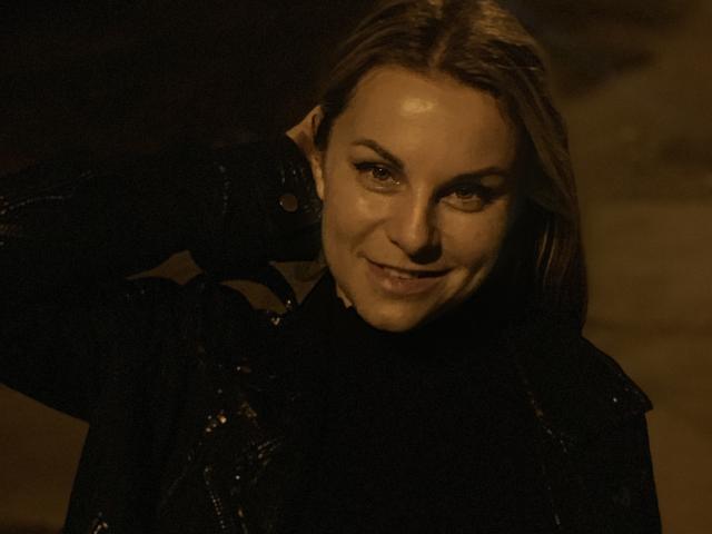 BelleLisaG with dirty blonde hair, smiling, wearing a black jacket, her shiny face lit warmly, hand behind her head, looking cute and slightly seductive.