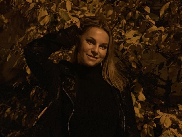 BelleLisaG con i capelli biondi, che indossa una maglia a collo alto nera e una giacca, sorridente, con una mano tra i capelli, illuminazione scura e suggestiva, circondata da foglie giallo-marrone..