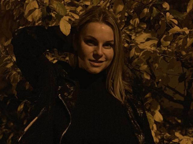 BelleLisaG, con i capelli biondi, indossa una maglia nera a collo alto e una giacca di pelle, sorride, ha una mano dietro la testa e si trova in mezzo a rami scuri e fogliosi, illuminata da una luce calda..