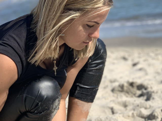 BelleLisaG con i capelli biondi, accovacciata su una spiaggia sabbiosa, indossa una maglietta nera e pantaloni di pelle nera, guarda in basso, con della sabbia sui pantaloni e una collana con una croce..