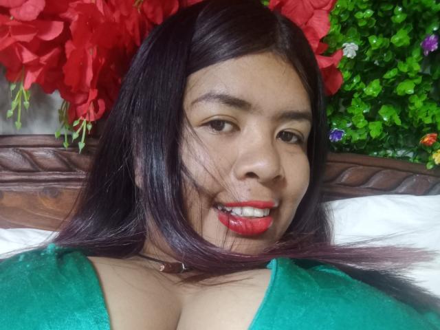 MariahSquirting69 with dark brown hair, red lipstick, green top, showing big tits, smiling, lying on bed with wooden headboard, red flowers, and green foliage in background.