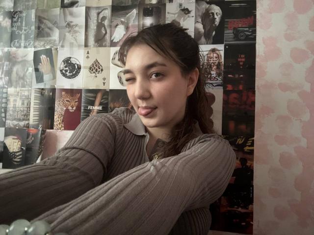 MichikoVivona, winking with a playful expression, wearing a ribbed, gray, long-sleeve shirt, has brown hair in a braid, and a cute nose ring, in a cozy room.