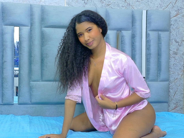 Tifannyy with curly black hair, pink satin robe open to reveal small tits, sitting on blue bed, right hand on robe, left hand on bed, smiling at camera.