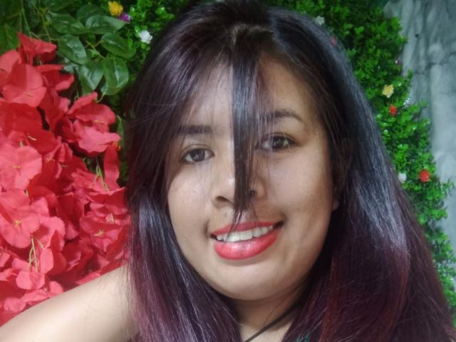 MariahSquirting69 has dark brown hair, red lipstick, and a cute smile, with red flowers on the left and green foliage on the right. She's looking at the camera.