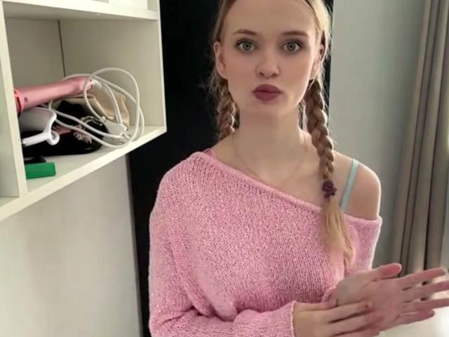 LawanaDavito with braided blonde hair, pink knit sweater, and blue strap, standing, hands slightly raised, pink hairdryer and white shelf in background.
