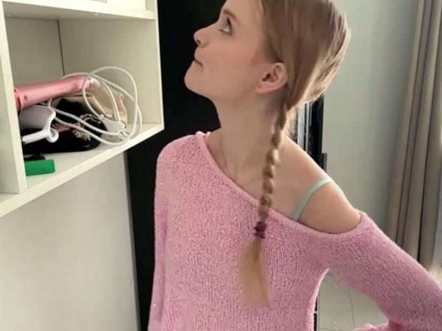 LawanaDavito with blonde braid wearing pink knit sweater, looking up at white shelf with pink hairdryer and white cables. Off-shoulder reveals blue strap.