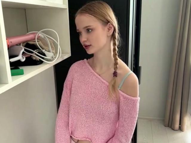 LawanaDavito with a side braid, wearing a pink, off-shoulder sweater, showing a blue bra strap, standing near a white shelf with a pink hairdryer.